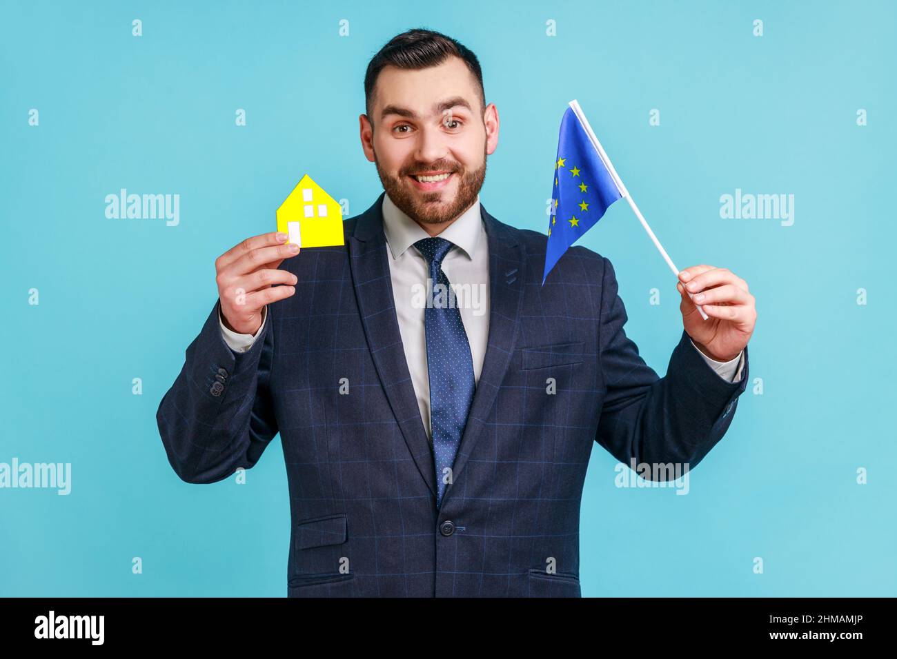 Gut aussehender Mann mit Bart, der einen Anzug im offiziellen Stil trägt, der das Modell des Papierhauses und die eu-Flagge hält und vom Leben in einem europäischen Land träumt. Innenaufnahme des Studios isoliert auf blauem Hintergrund. Stockfoto