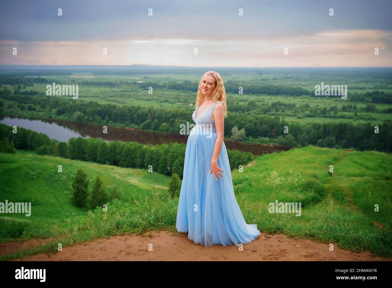 Schwangere in blauem Kleid, grünen Hügeln, Fluss und Himmel Stockfoto
