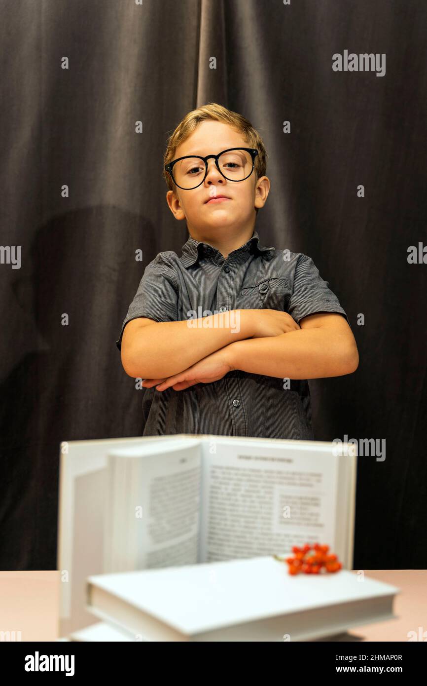 Ein Junge mit Brille faltete seine Arme auf seiner Brust, ein ernsthafter Blick, ein Stapel Bücher, zurück zur Schule, Lernschwierigkeiten Stockfoto