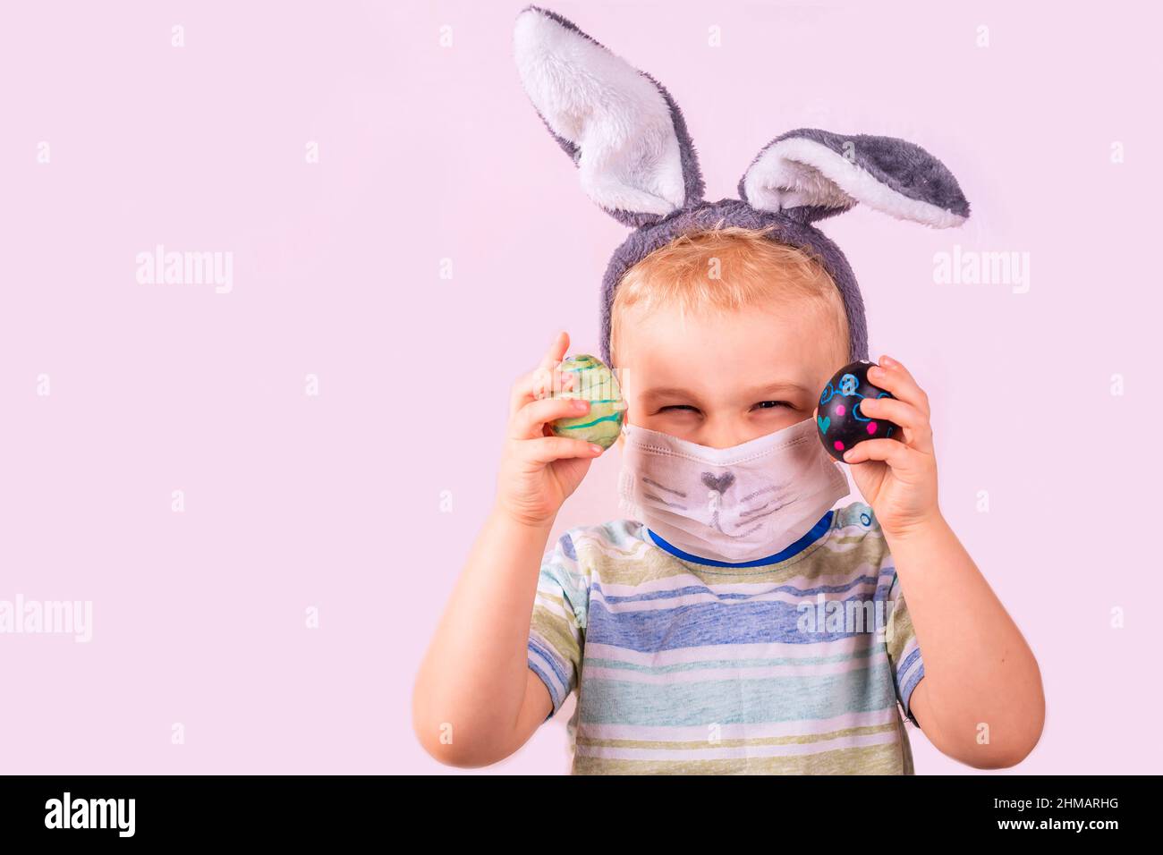 Niedlichen Baby Junge in Kaninchen Hase Ohren auf dem Kopf und Schutzmaske mit farbigen Eiern auf rosa Hintergrund. Fröhlich lächelndes glückliches Kind. Kovid Ostern holida Stockfoto