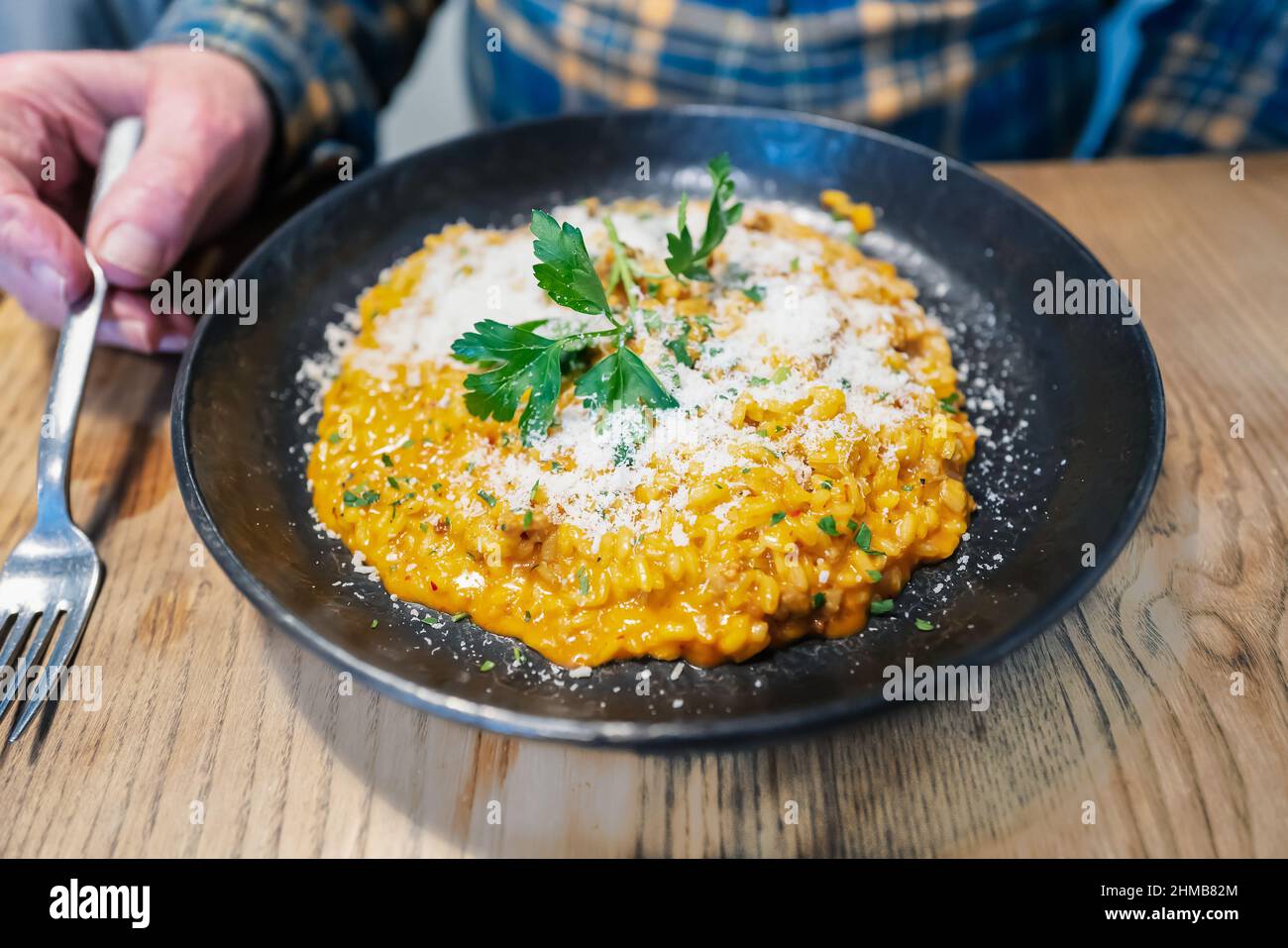 Risotto in einem großen Gericht mit Parmesankäse. Ein Mann sitzt an einem rustikalen Holztisch und hält eine Gabel. Stockfoto
