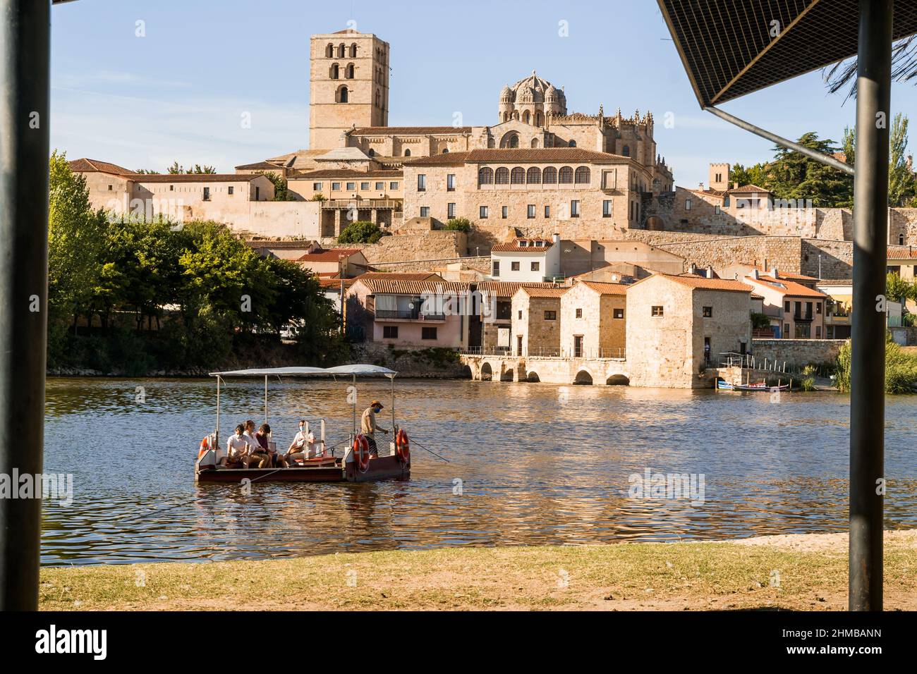 Blick auf die Kathedrale mit den Mühlen und den Fluss Duero, wo sich an einem sonnigen Sommertag in Zamora ein Lastkahn mit Touristen über das Wasser zieht, Stockfoto