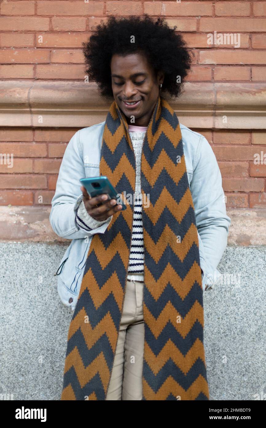 Lächelnder afroamerikanischer Mann mit Afro-Frisur und legerer Kleidung, der im Freien mit dem Mobiltelefon telefoniert. Stockfoto