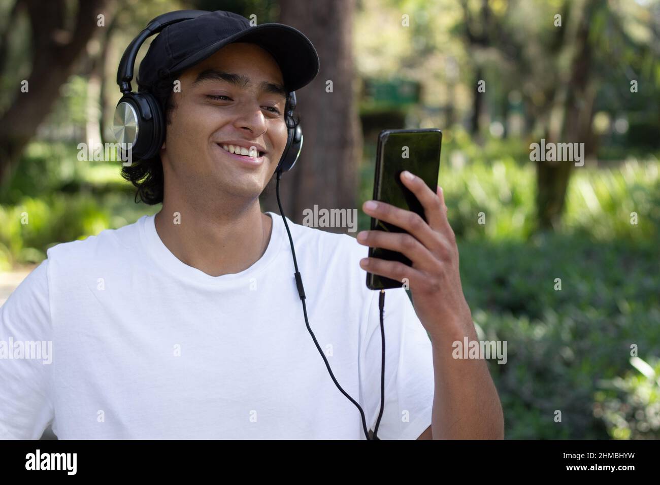 Der junge Mann hörte Musik mit Kopfhörern, während er lächelnd und entspannt mitten in der Natur durch den Park schlendert, mit einer Mütze und einem weißen t Stockfoto