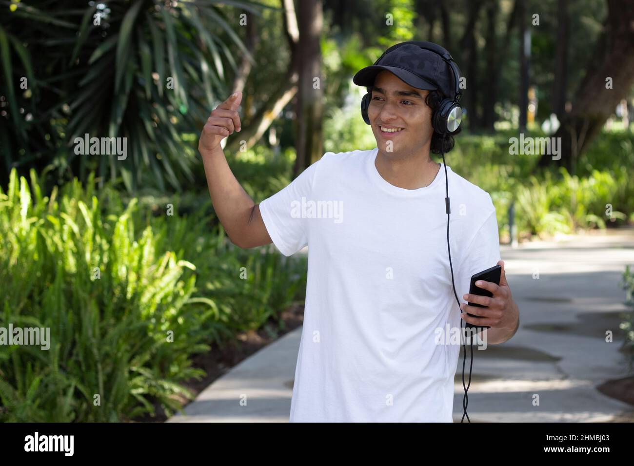 Der junge Mann hörte Musik mit Kopfhörern, während er lächelnd und entspannt mitten in der Natur durch den Park schlendert, mit einer Mütze und einem weißen t Stockfoto
