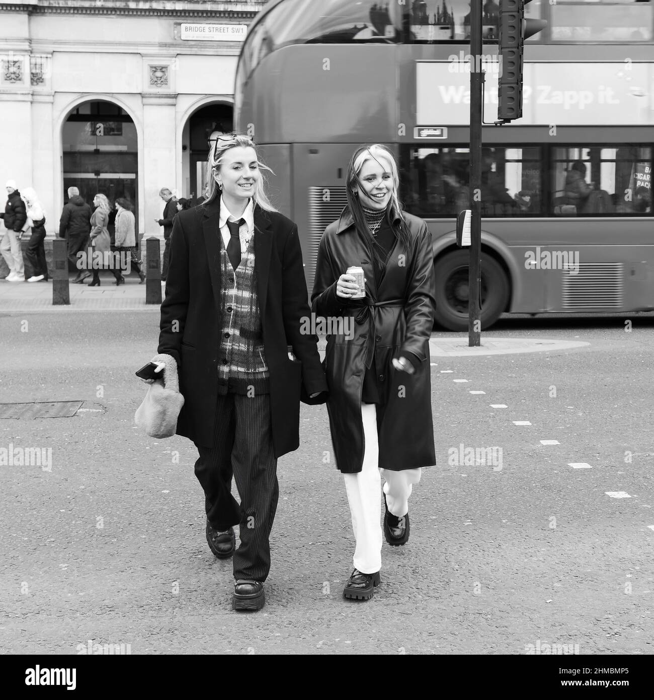 London, Greater London, England, Februar 05 2022: Zwei trendige Frauen, eine in einer Weste, lächeln und schmunzeln mit einem Bus über die Bridge Street Stockfoto