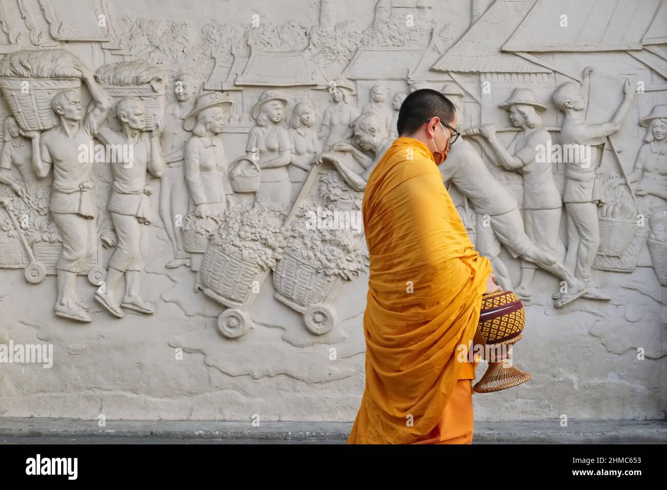 Ein buddhistischer Mönch, der eine Almosenschale trägt, passiert an einem Fries außerhalb der Yodpiman Flower City (Markt), Bangkok, Thailand, und zeigt eine geschäftige Marktszene von früher Stockfoto