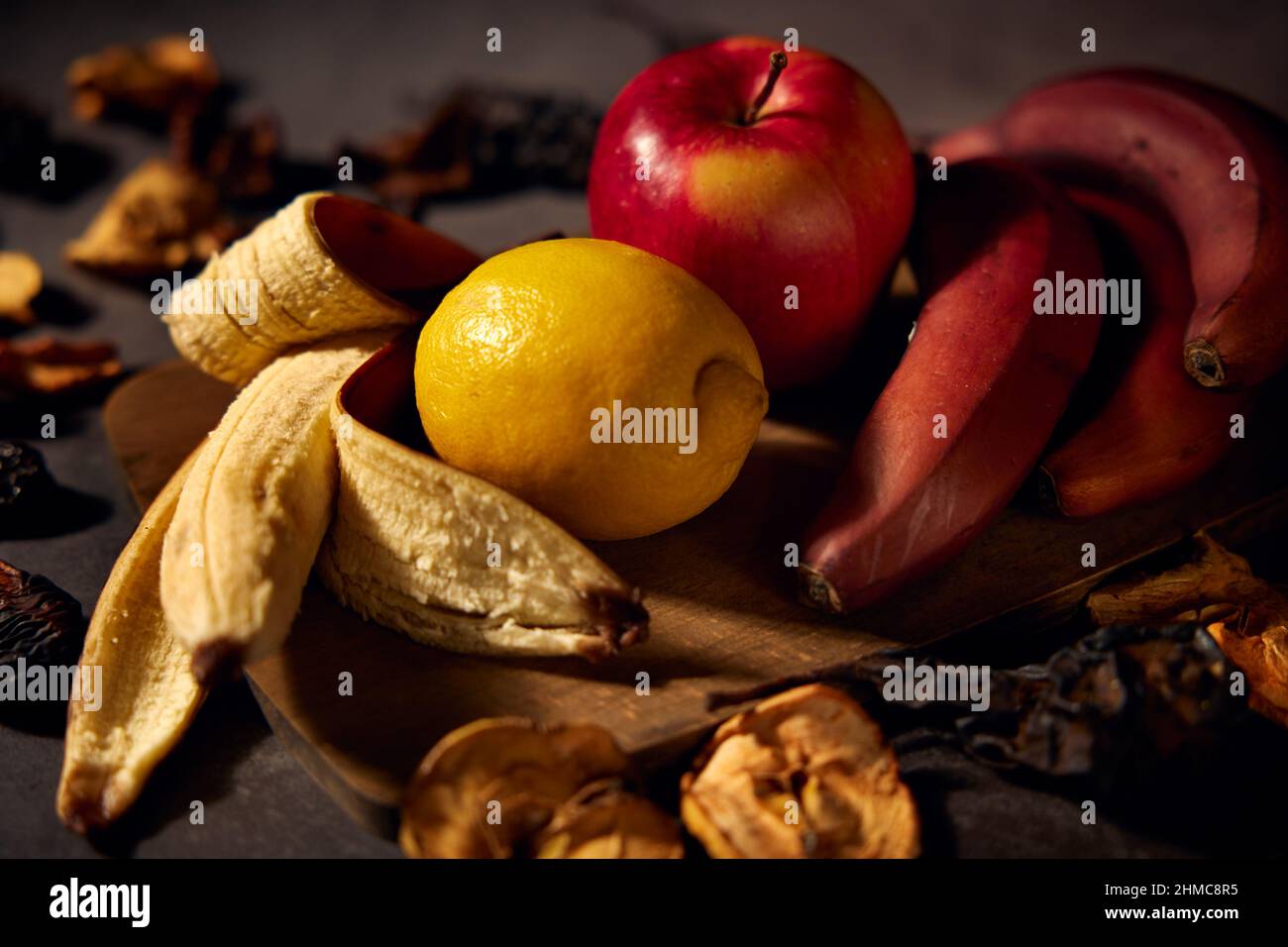 Obsttafel. Bananen, Zitrone, Apfel und getrocknete Früchte in Scheiben geschnitten auf einem Holzbrett mit einem dunklen verschwommenen Hintergrund. Stockfoto