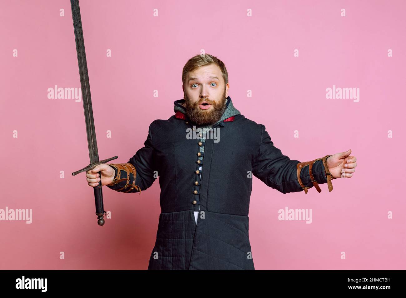 Ein junger schockierter Mann, ein mittelalterlicher Krieger oder Bogenschütze mit Schwert, der mit offenem Mund auf einem rosafarbenen Studiohintergrund die Kamera ansah. Vergleich von Stockfoto