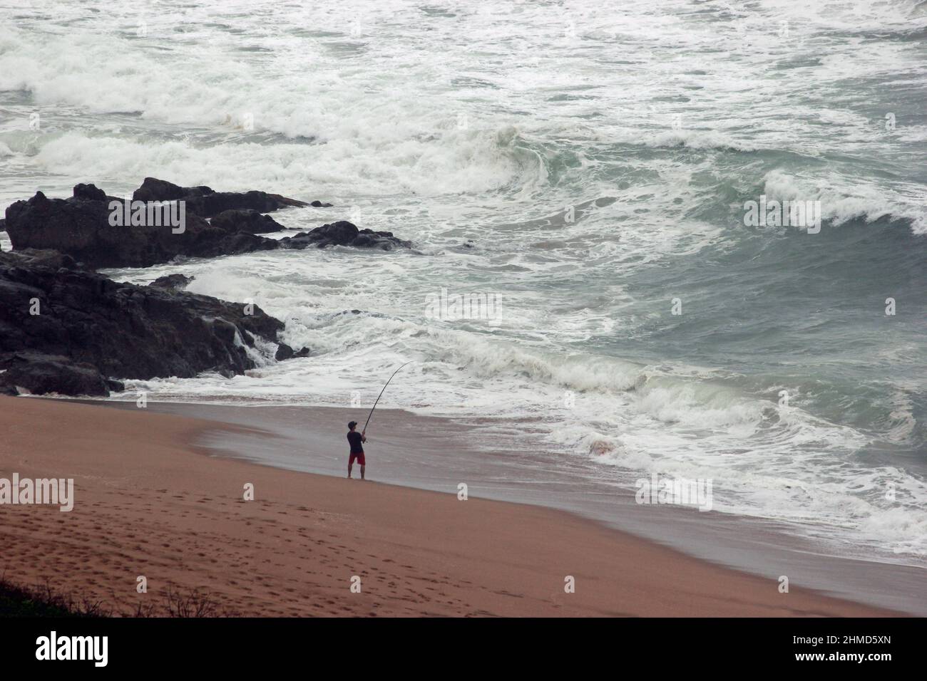 Junge Angler, der am Strand mit Rute in den Händen stand und versuchte, Fische zu fangen, dunkle Felsen auf seiner linken Seite und weiße Wellen vor ihm. Stockfoto