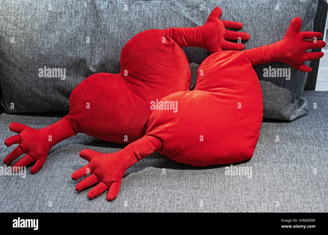 Zwei rote Plüschkissen in Herzform mit Händen auf einem grauen Sofa. Stockfoto