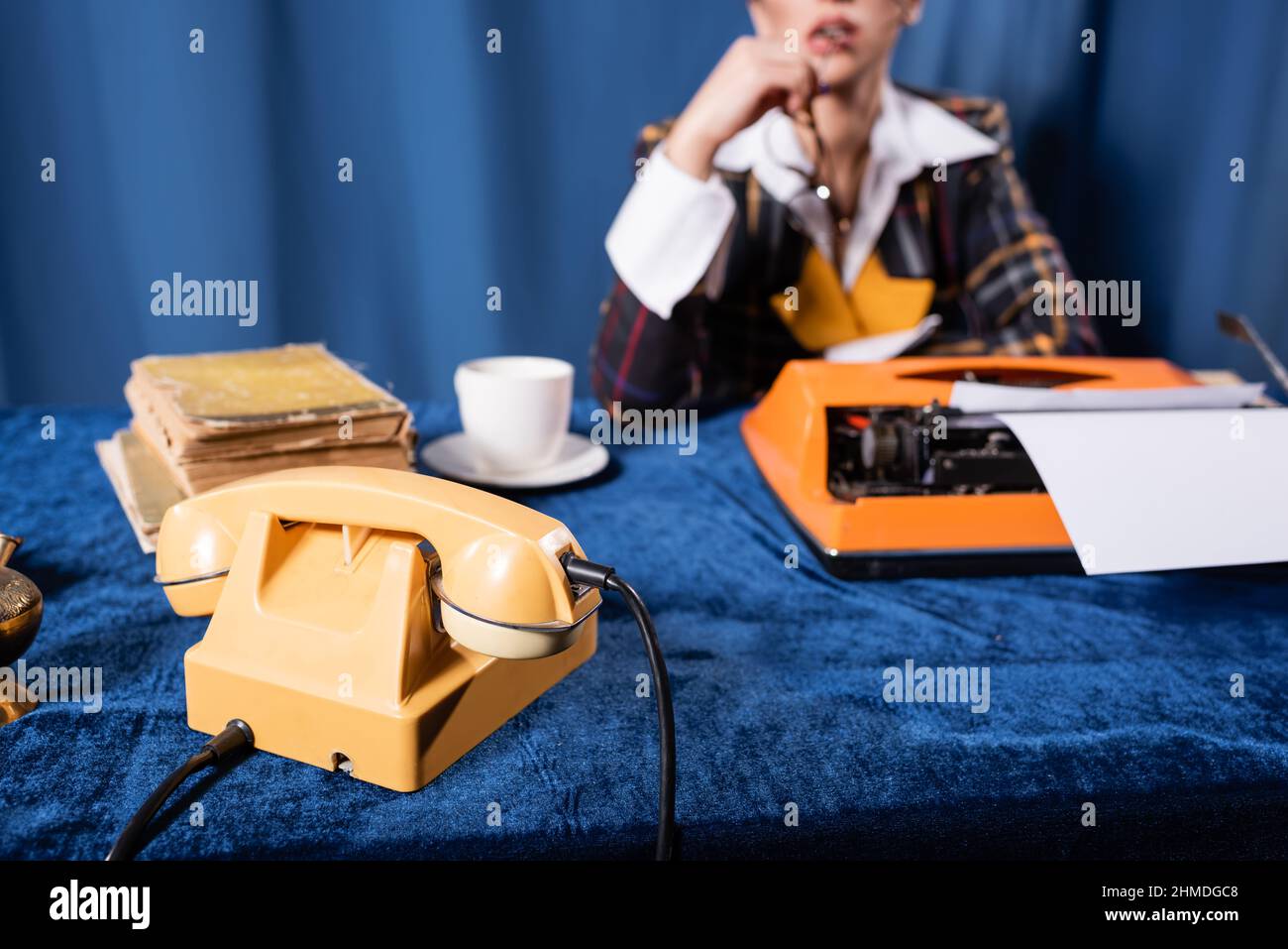 Teilansicht einer verschwommenen Frau in der Nähe eines Vintage-Telefons, einer Schreibmaschine und Büchern auf einer blauen Velours-Tischdecke Stockfoto
