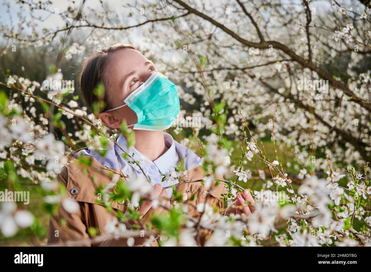 Eine allergische Frau, die im Frühling an einer saisonalen Allergie leidet, im Frühling im blühenden Garten posiert und unter blühenden Bäumen eine medizinische Maske trägt. Frühjahrsallergiekonzept Stockfoto
