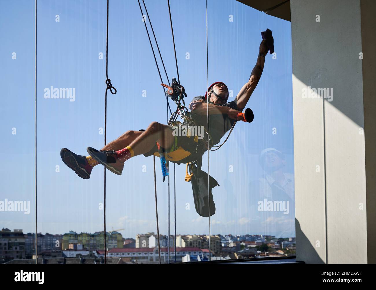 Arbeiter für den industriellen Bergsport, der am Kletterseil hängt und an den Reinigungsfenstern des Hochhauses hängt. Mann, der Sicherheitsausrüstung verwendet, während er das Fenster des Wolkenkratzers mit einem Lappen abwischt. Stockfoto