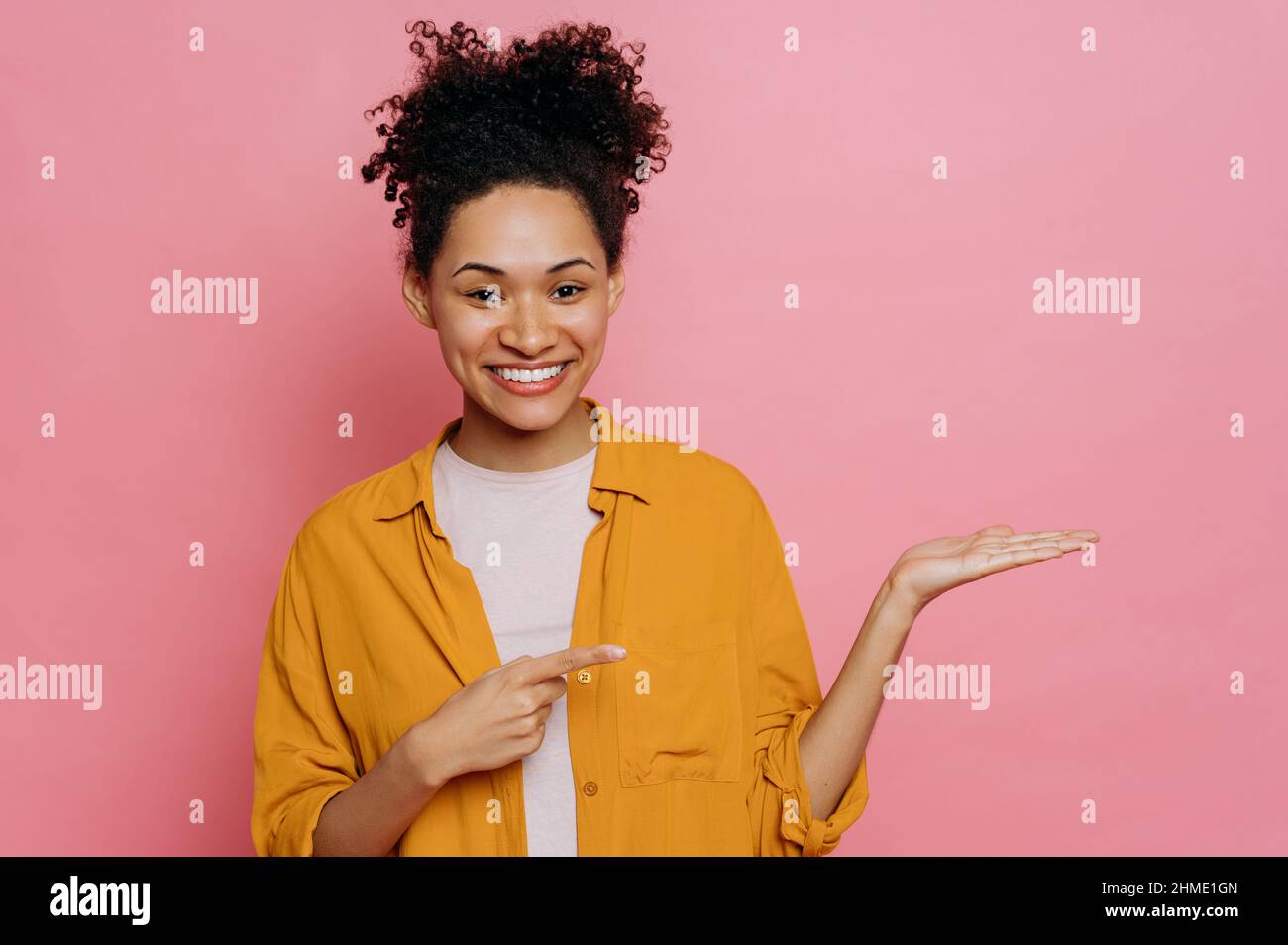 Modell für die Präsentation. Freundliche positive afroamerikanische Mädchen mit lockigem Haar, zeigt mit den Händen auf leeren Raum, Werbefläche, stehen auf isolierten rosa Hintergrund, schaut auf die Kamera, lächeln Stockfoto