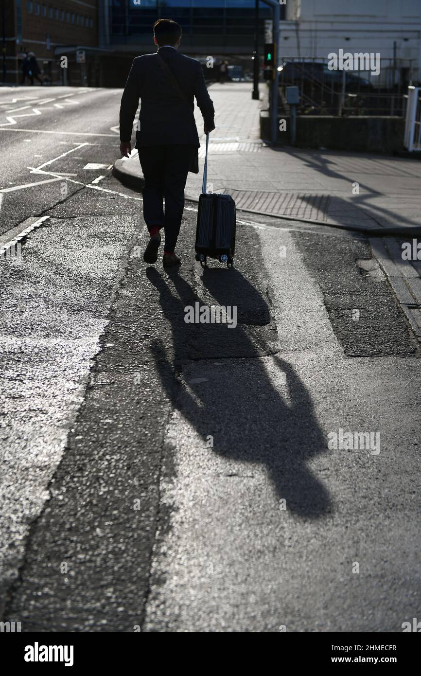 Mögliches Konzeptbild, eine Silhouette eines Mannes in einem Anzug, der mit einem Rollkoffer-Trolley mit einem perfekten Schatten seiner Linie hinter dem Bürgersteig läuft. Stockfoto