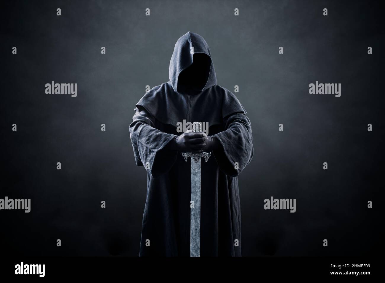 Krieger mit Kapuzenumhang und mittelalterlichem Schwert auf dunklem nebligen Hintergrund Stockfoto