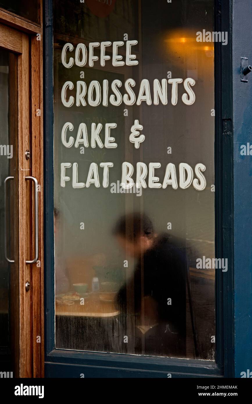 Männlicher Kunde sitzt in einem Café mit einem verblendeten Fenster, das Kaffee, Croissants, Kuchen und Fladenbrot anwirbt. Edinburgh, Schottland, Großbritannien. Stockfoto
