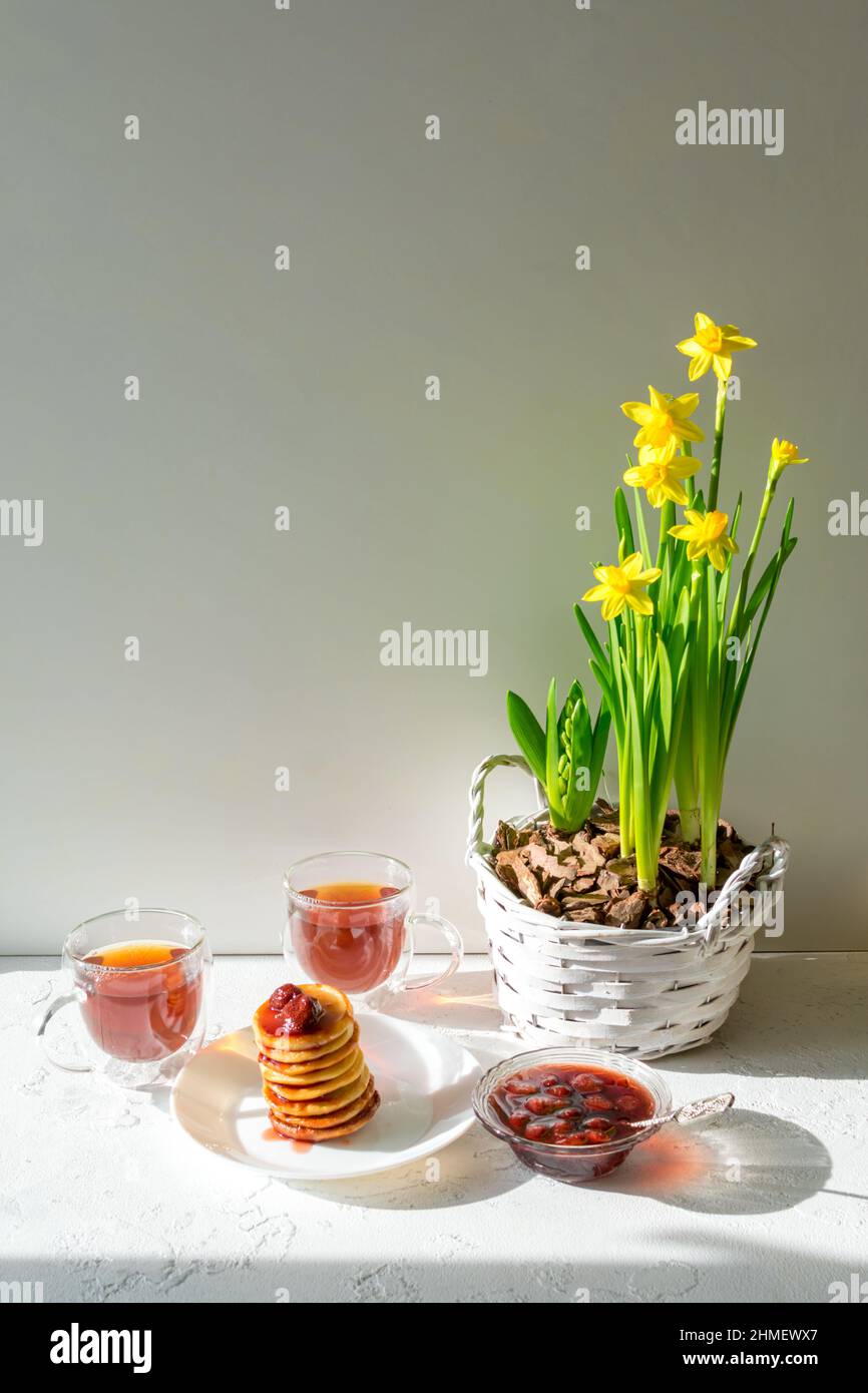 Krapfen mit Erdbeermarmelade, zwei Tassen Tee und ein Bouquet von gelben Narzissen in einem Korb auf weißem Hintergrund. Stockfoto