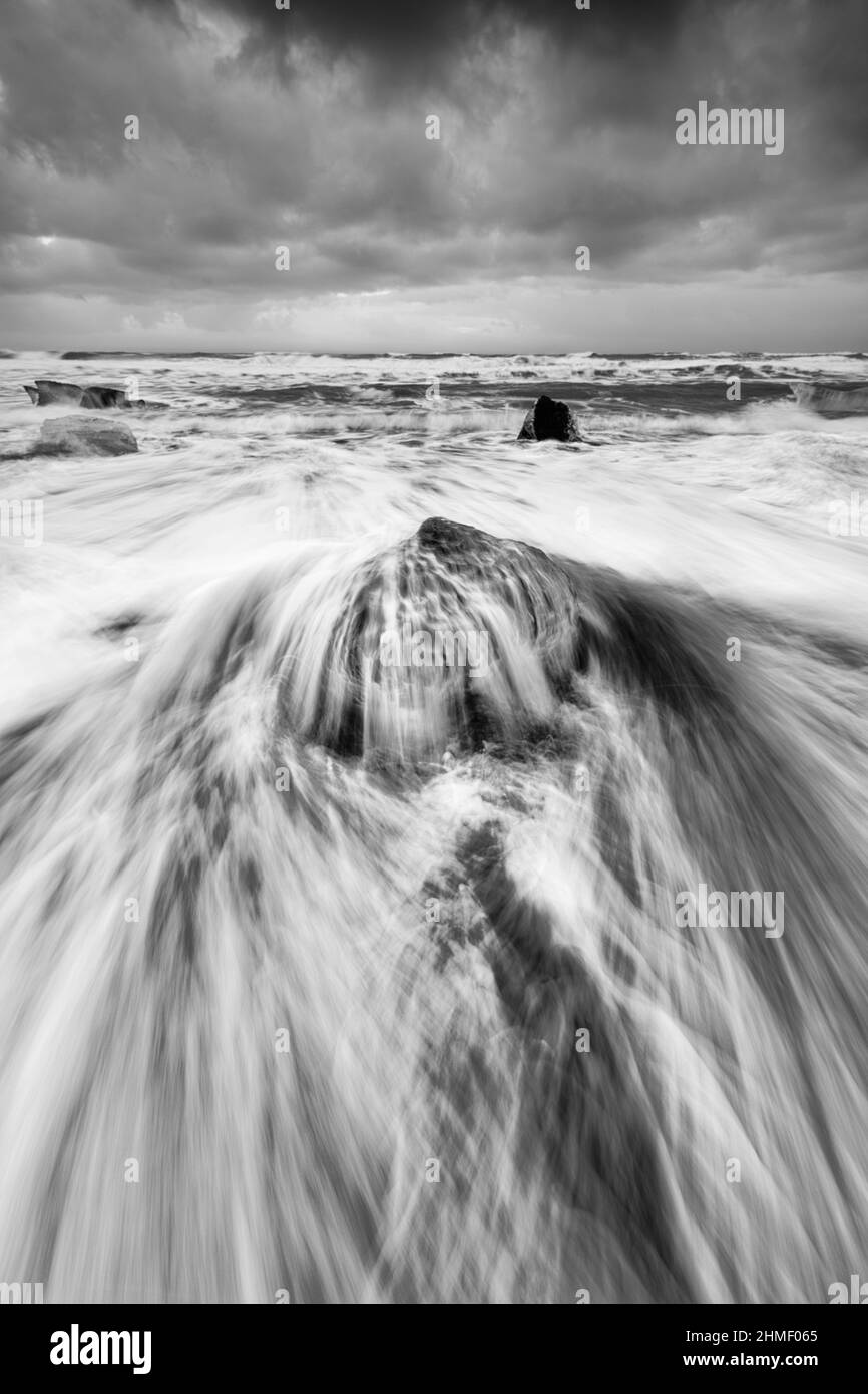 Ein Eisblock wird von einer Welle auf einem Strand schwarz mit starker Brandung überspült, die Wasserbewegung ist zu sehen (lange Exposition, klare Spuren der Bewegung), a Stockfoto