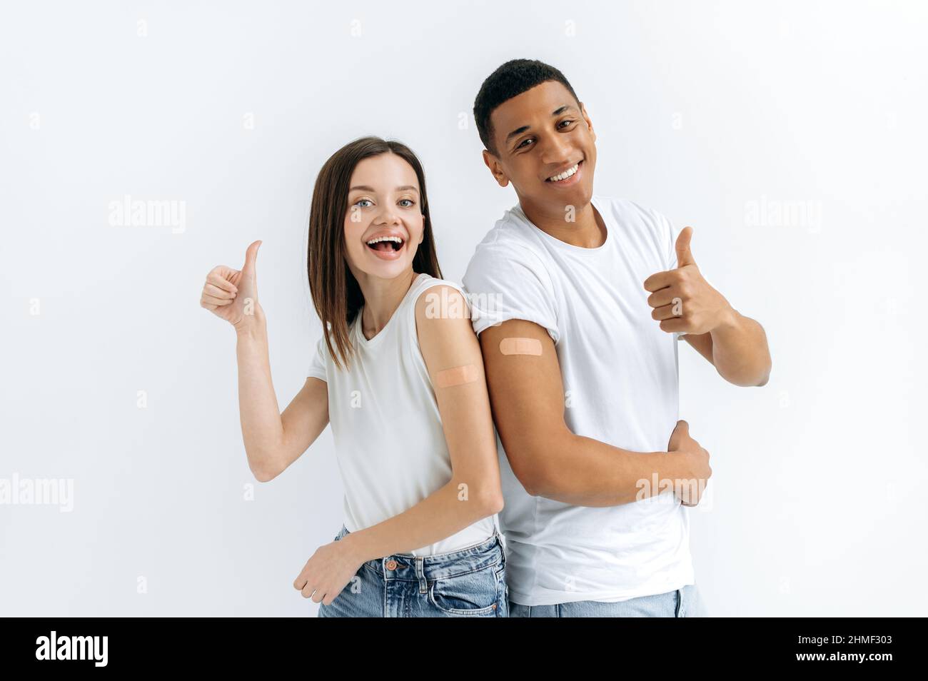 Fröhliches glückliches kaukasisches Mädchen und hispanischer Kerl, sind geimpft, stehen auf weißem Hintergrund mit medizinischen Pflaster auf ihren Schultern, schauen auf die Kamera, lächelnd, zeigen Daumen nach oben Geste Stockfoto