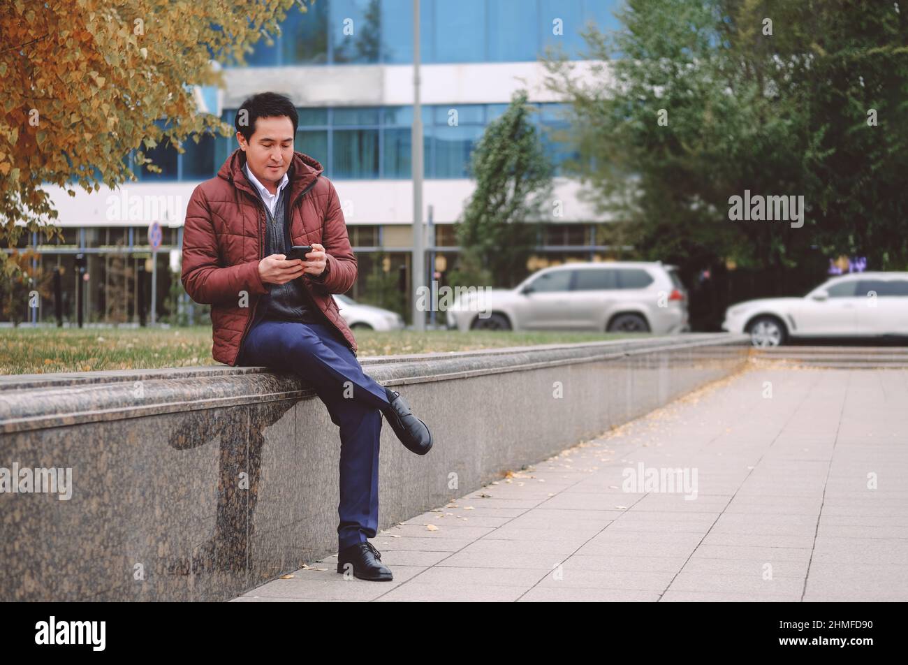 Ein asiatischer Mann mittleren Alters, der mit dem Telefon auf der Bank sitzt. Stockfoto