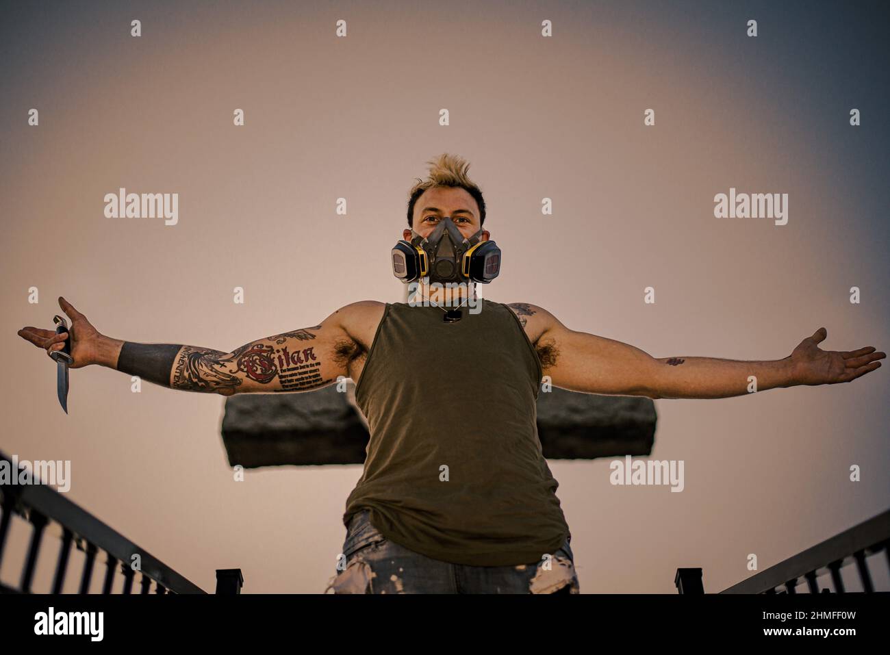 Junger Punk-Mann mit Tattoos, Maske und Messer, ausgestreckte Arme, Blick aus dem unteren Winkel Stockfoto