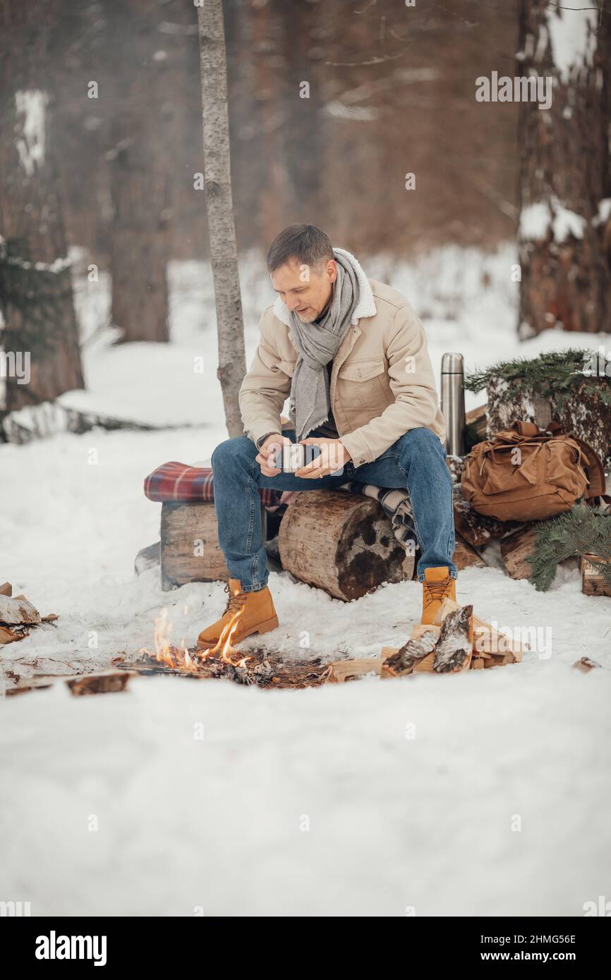 Ein brutaler Mann im Winterwald am Feuer, der Kaffee trinkt Stockfoto