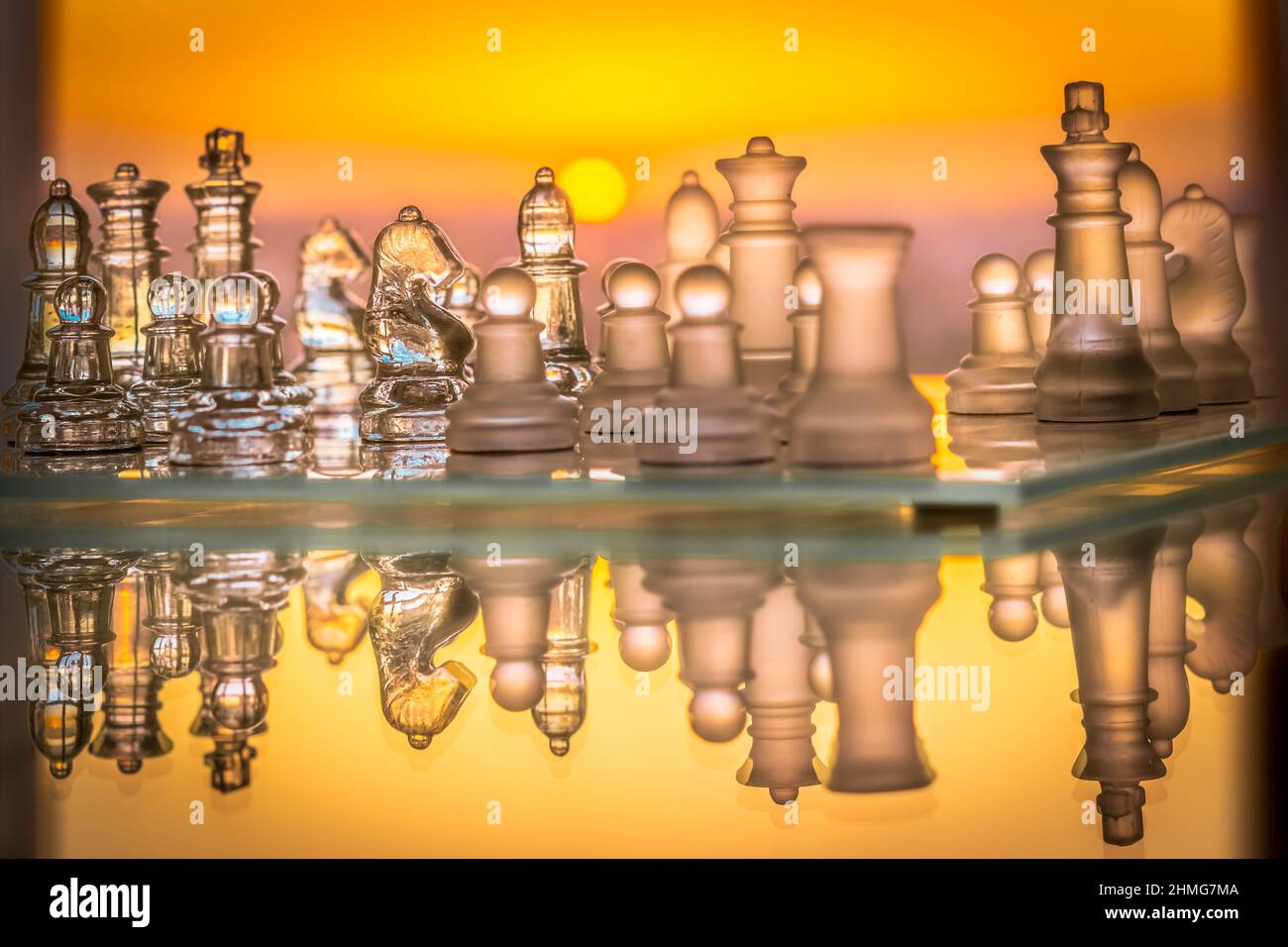 Schachspiel mit Glasmaterialstücken und Schachbrett horizontal reflektiert mit einem sattorange farbigen Sonnenuntergang im Hintergrund. Stockfoto