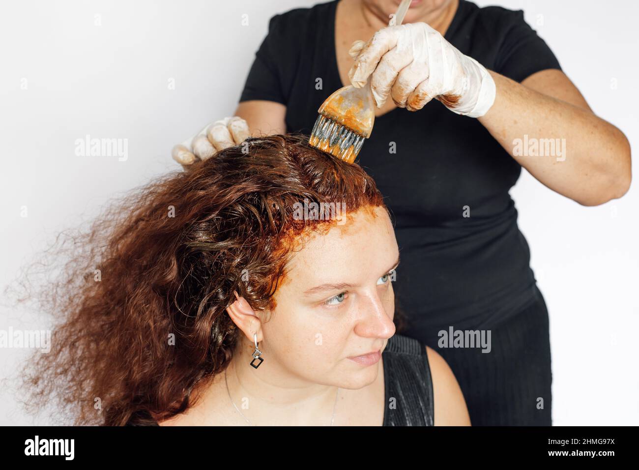 Ältere Dame hinter Frau färbt Haare in rot mit Pinsel in den Händen mit Latexhandschuhen auf, weißer Hintergrund. Haarfärbung zu Hause. Graue Farbe wird loswerden Stockfoto