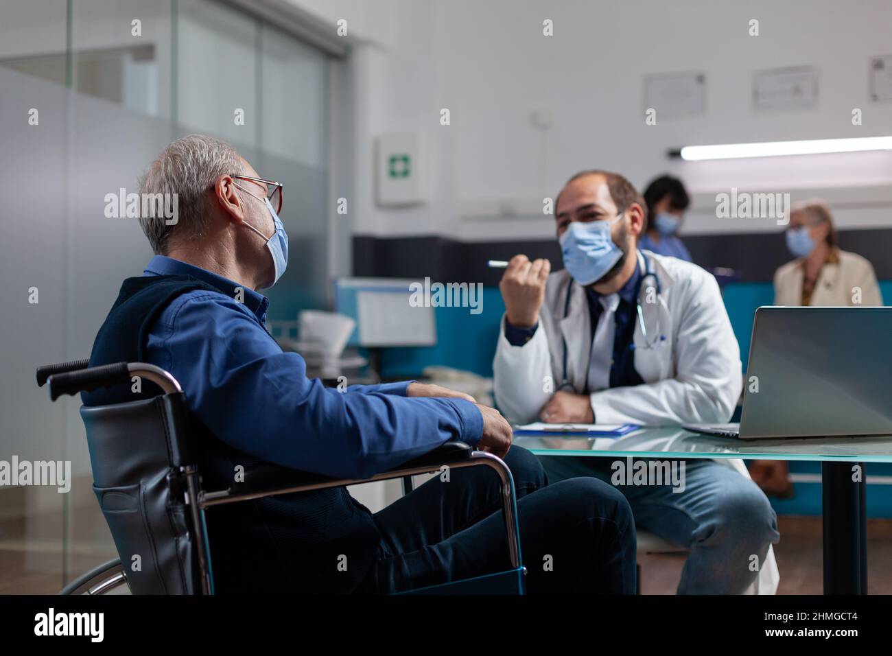 Rentner, der während einer Coronavirus-Pandemie im Rollstuhl sitzt, bei einem Kontrollbesuch mit einem Sanitäter. Arzt, der Gesundheitskonsultation macht, im Gespräch mit einem pensionierten Patienten mit körperlicher Behinderung im Büro. Stockfoto