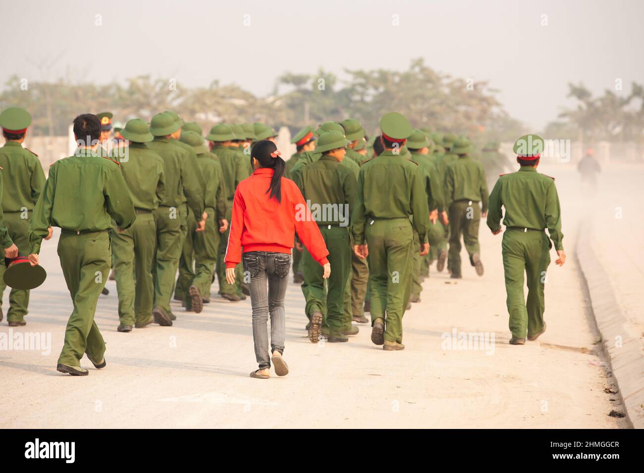 Rückansicht eines Teenagers im roten Hemd, das mit einer Gruppe junger vietnamesischer Soldaten auf einer Straße, vietnamesischen Militärakademien, läuft. Vietnam. Stockfoto
