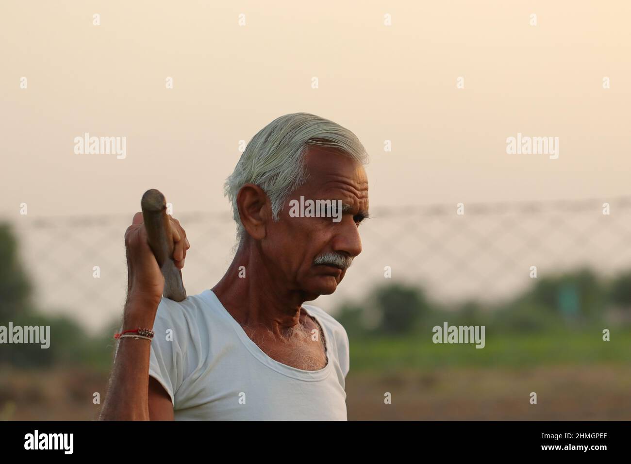 Nahaufnahme eines älteren indischen Bauern, der bei Sonnenuntergang mit einer Schaufel auf der Schulter und einer weißen Weste auf dem Feld steht Stockfoto