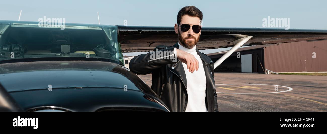 Bärtiger Pilot in Lederjacke und Sonnenbrille in der Nähe des Hubschraubers stehend, Banner Stockfoto