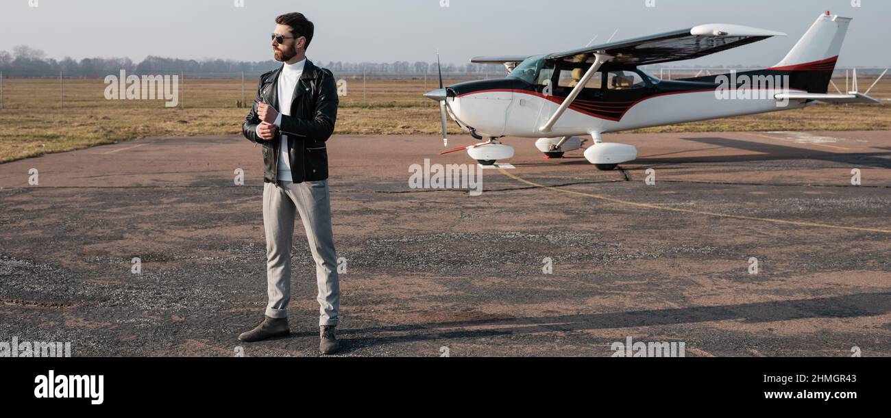 Die ganze Länge des stilvollen Piloten in Lederjacke und Sonnenbrille in der Nähe des Hubschraubers stehend, Banner Stockfoto