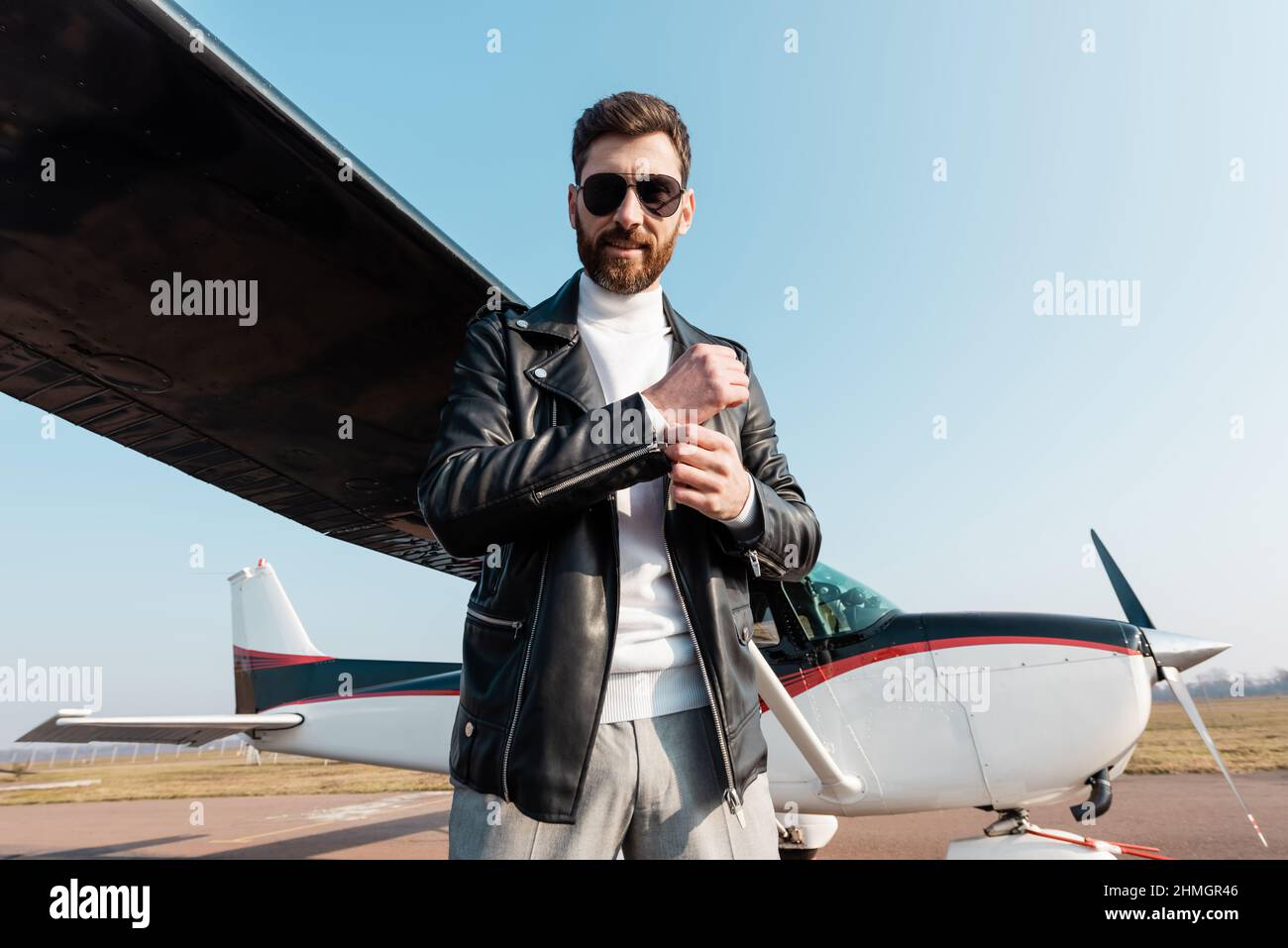 Fröhlicher Pilot in Sonnenbrille und Lederjacke, der in der Nähe des Flugzeugs steht Stockfoto