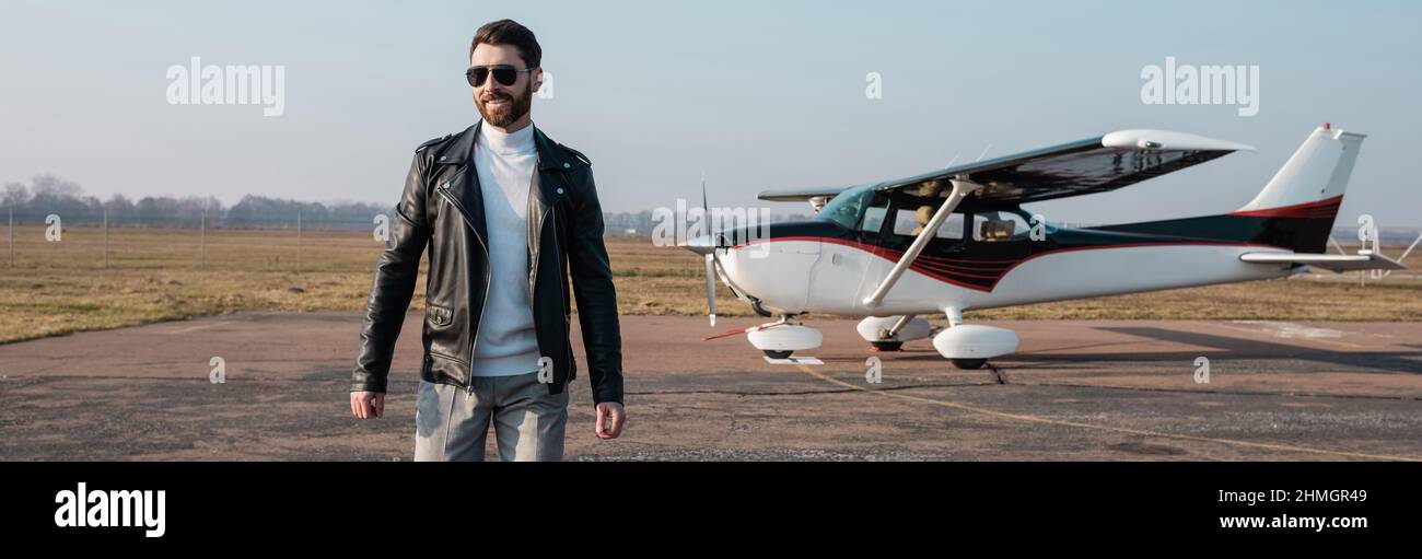 Glücklicher Pilot in stilvoller Lederjacke und Sonnenbrille, der in der Nähe des Hubschraubers, Banner, läuft Stockfoto