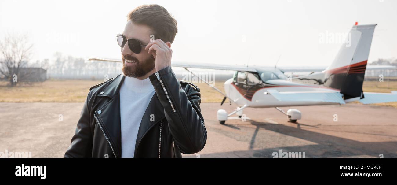 Zufrieden Pilot in stilvoller Lederjacke Anpassung Sonnenbrille in der Nähe Hubschrauber, Banner Stockfoto