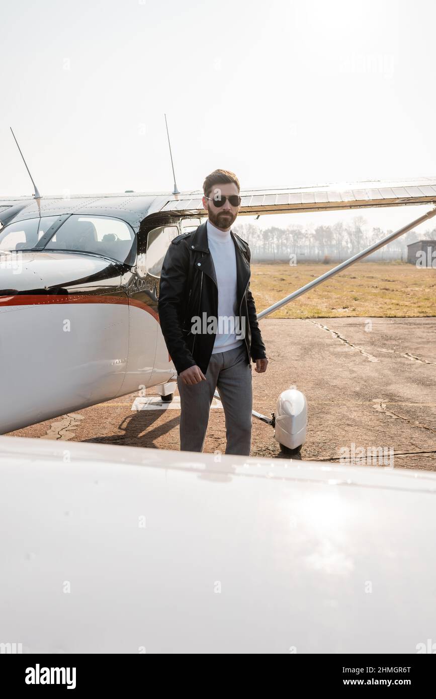 Bärtiger Pilot in schwarzer Lederjacke und Sonnenbrille in der Nähe des Hubschraubers Stockfoto