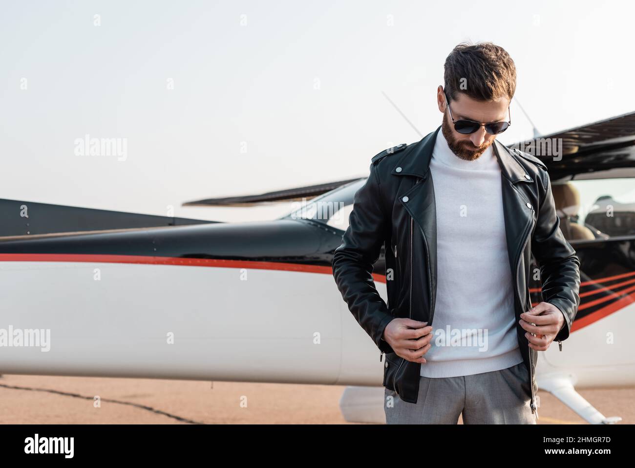 Bärtiger Pilot in stilvoller Sonnenbrille mit verstellter Lederjacke in der Nähe des Flugzeugs Stockfoto