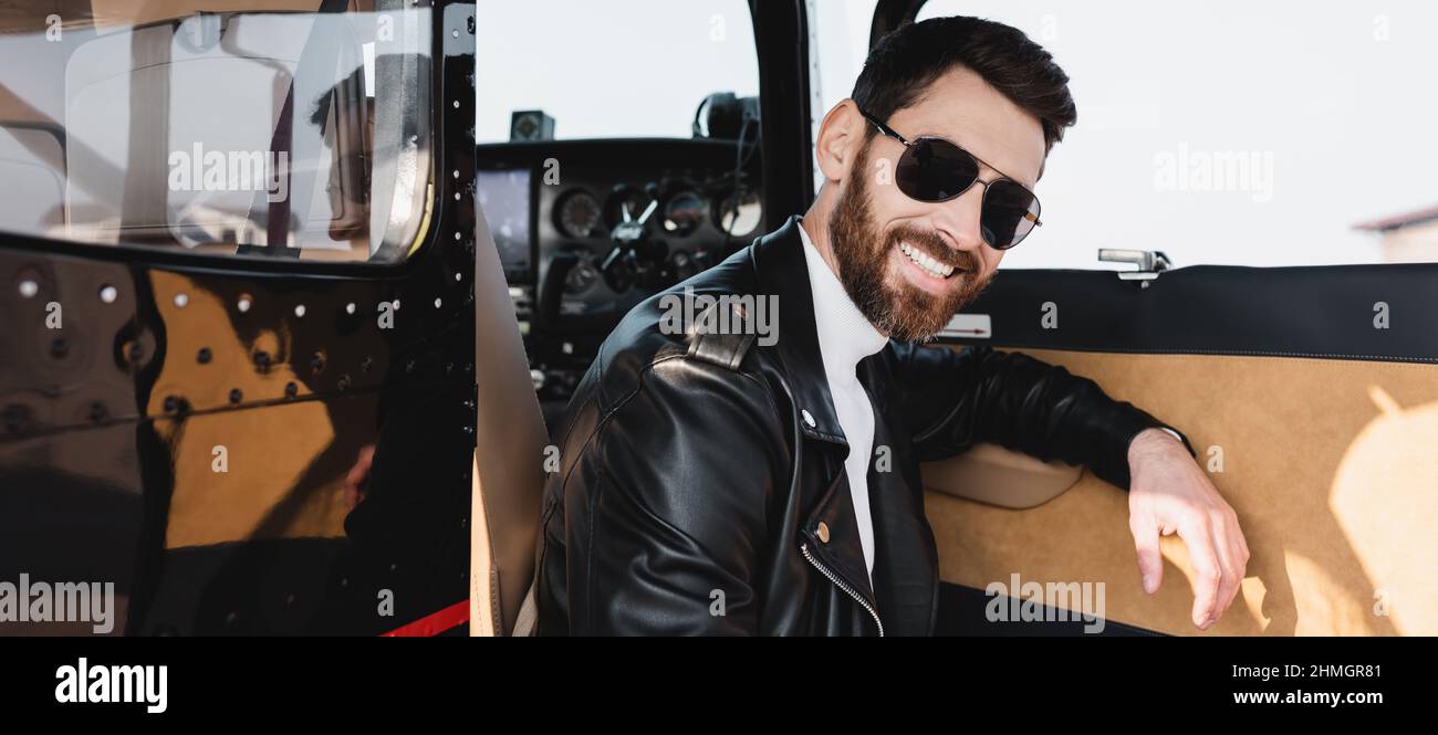 Glücklicher Pilot in stilvoller Sonnenbrille und Lederjacke in der Nähe der geöffneten Tür des Hubschraubers, Banner Stockfoto