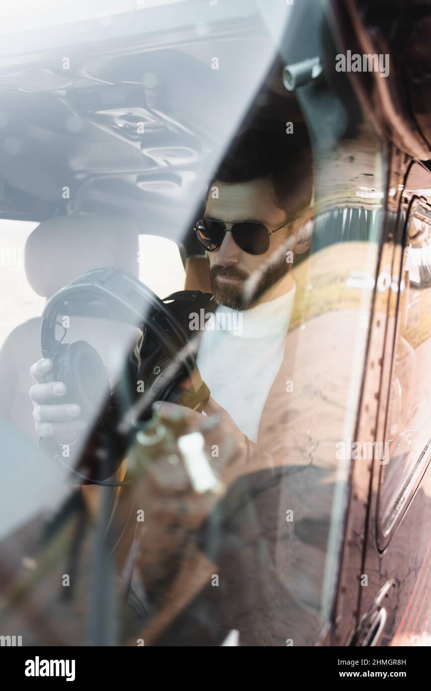 Blick auf den bärtigen Piloten in einer stilvollen Sonnenbrille, die Kopfhörer im Cockpit durch die Windschutzscheibe des Hubschraubers hält Stockfoto