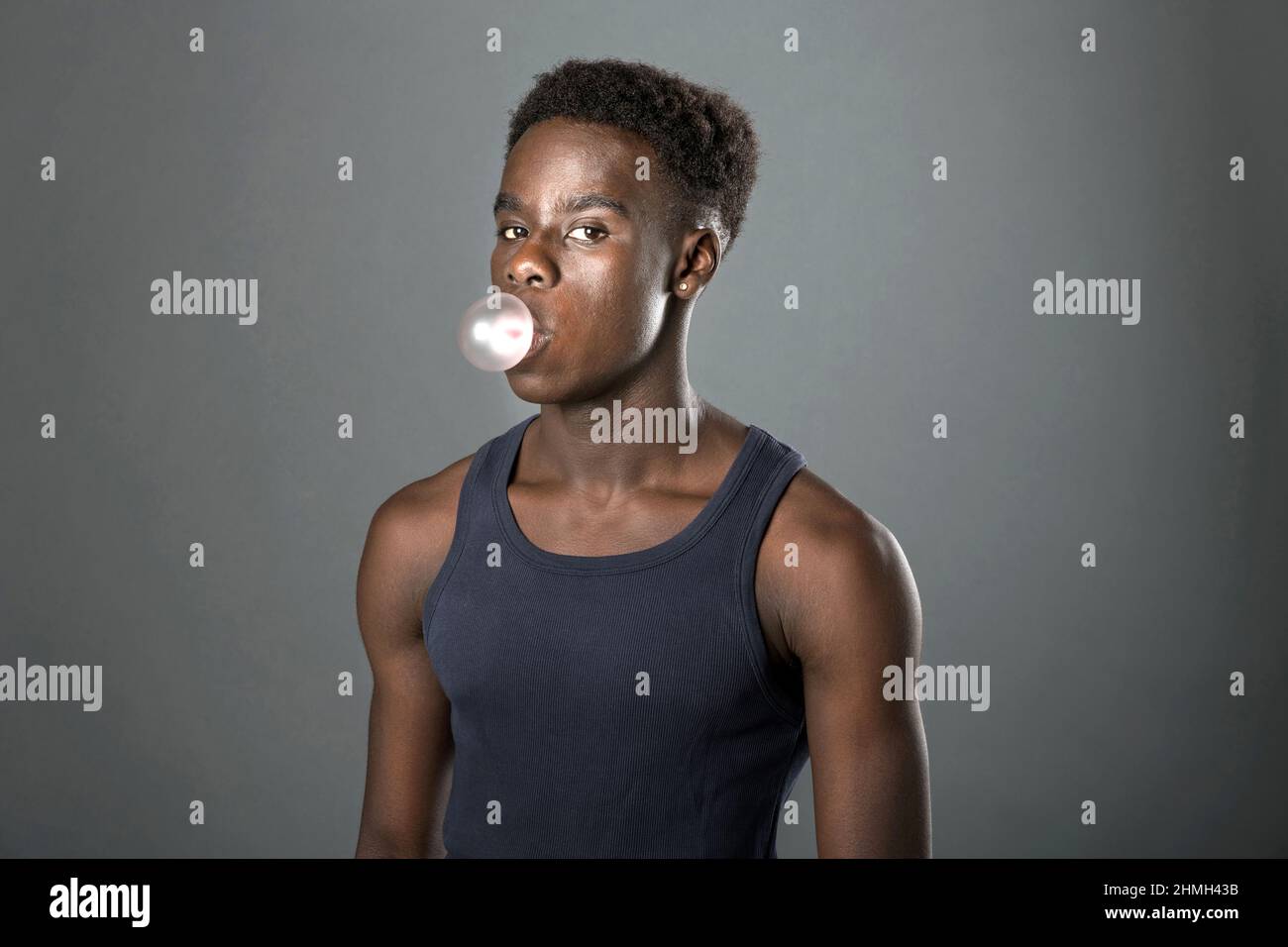 Der junge schwarze Kerl im ärmellosen Oberteil posiert über einem grauen Studiohintergrund und bläst Blasen mit Kaugummi, während er seitlich auf die Kamera sieht Stockfoto