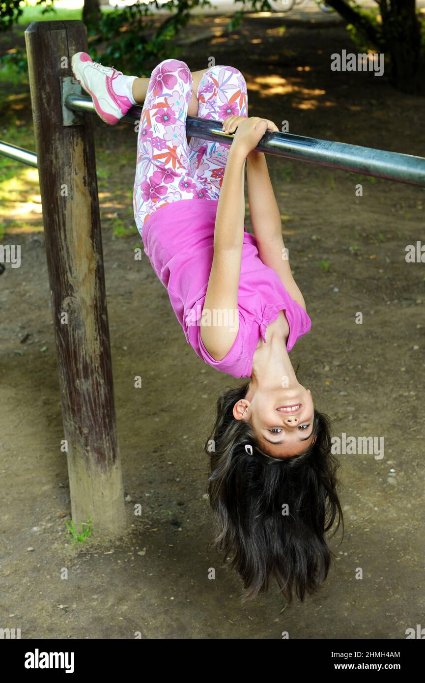 Freundliches, agiles kleines Mädchen, das kopfüber an einer Bar auf einem Geländer im Freien hängt, mit ihrem langen Haar, das auf den Boden fällt und einem glücklichen Lächeln Stockfoto