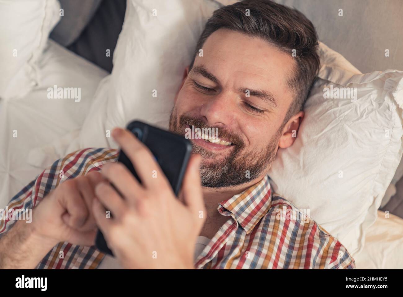 Süchtiger kaukasischer Mann mittleren Alters mit Gesichtshaar, das im Bett lag und den ganzen Tag über Social Media scrollte. Hochwertige Fotos Stockfoto