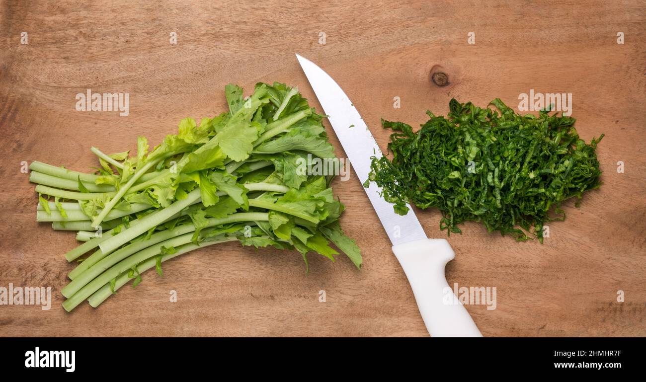 Gehackte Rettich-Grüns oder Blätter für Salat, essbare und gesunde Gemüse-Grüns auf einem hölzernen Hintergrund mit einem Messer, von oben genommen Stockfoto