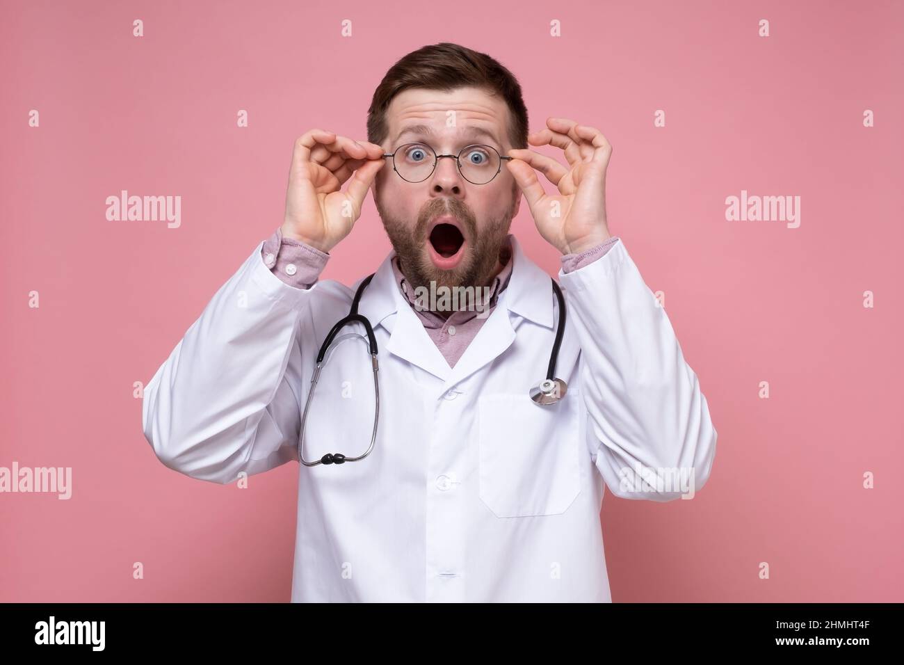Junger Arzt im weißen Mantel, mit Stethoskop um den Hals, geöffnetem Mund bei Schock und sieht mit großen Augen, die eine Brille halten. Stockfoto