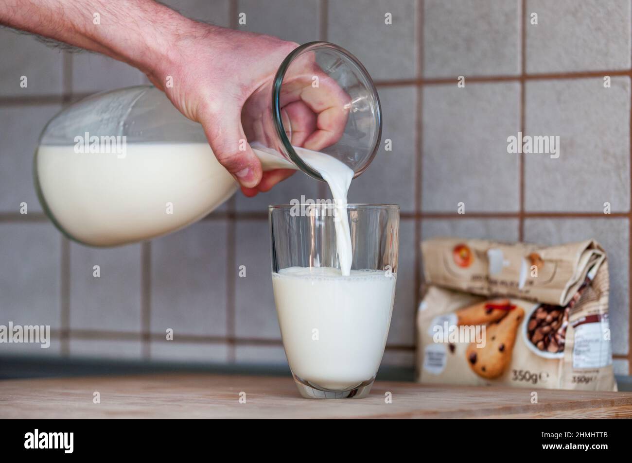 Die Hand eines Mannes hält einen Krug und gießt Milch in ein Glas. Eine Packung Kekse verschwimmt im Hintergrund Stockfoto