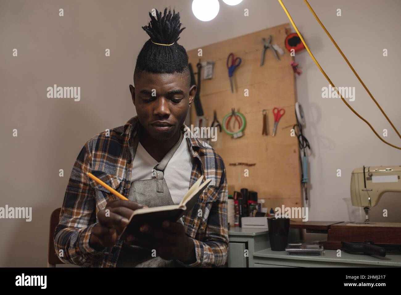 afroamerikanischer junger Handwerker mit Dreadlocks, der in einem Buch schrieb, während er in einer Lederwerkstatt arbeitete Stockfoto