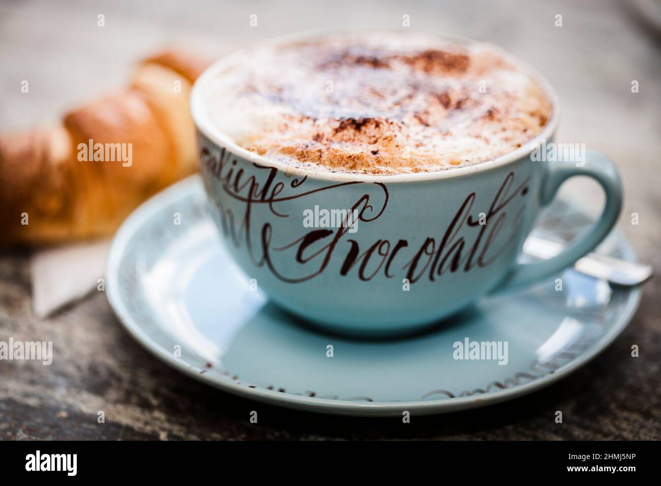 Heiße Schokolade mit einem Prise Kakaopulver in einem dekorativen Becher mit einem Croissant auf einer rustikalen Holzarbeitsfläche Stockfoto